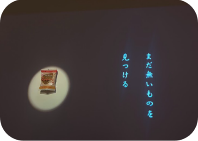 暗い壁にスポットライトで照らされたチキンラーメンと「まだ無いものを見つける」の文字