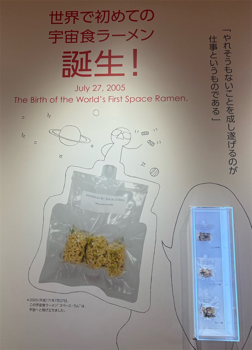 「世界で初めての宇宙食ラーメン誕生！」と書かれた展示パネルと宇宙食ラーメンの実物